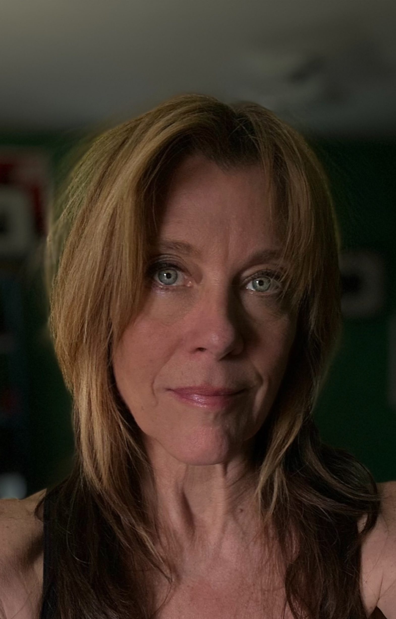 A woman with long brown hair wearing a black tank top looks at the camera, in front of a green wall with 2 framed pictures...